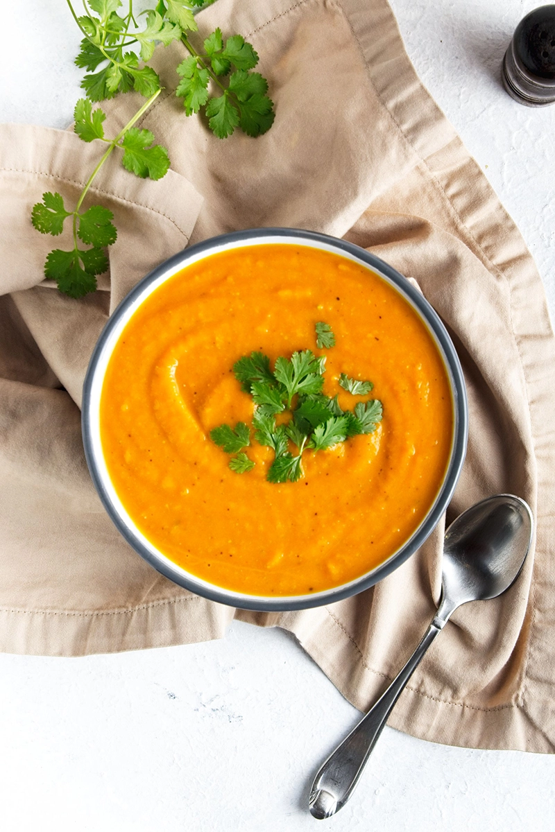 A bowl of sweet potato miso soup garnished with fresh cilantro sits on a beige cloth napkin with a spoon beside it.