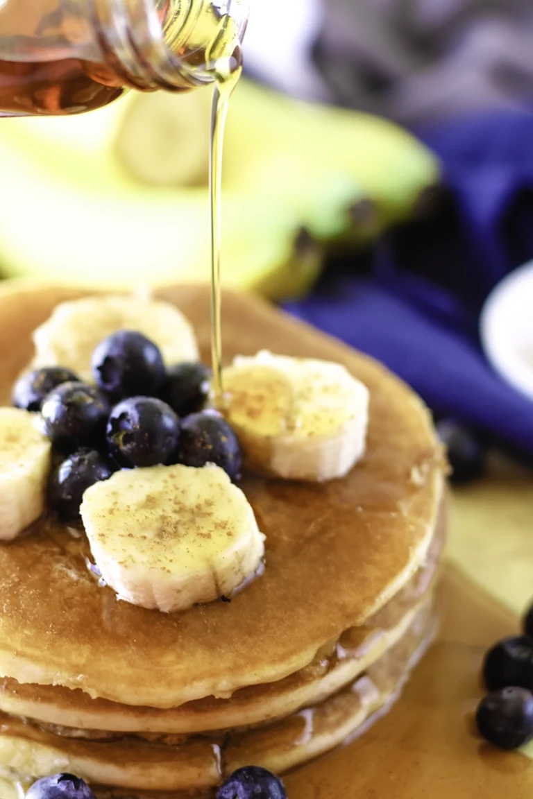 A stack of pancakes with blueberries and bananas being drizzled with syrup.