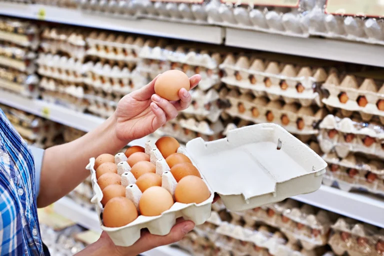 A person holds an egg carton with an open slot in a grocery store aisle, contemplating which egg substitute for breakfast best fits their needs.