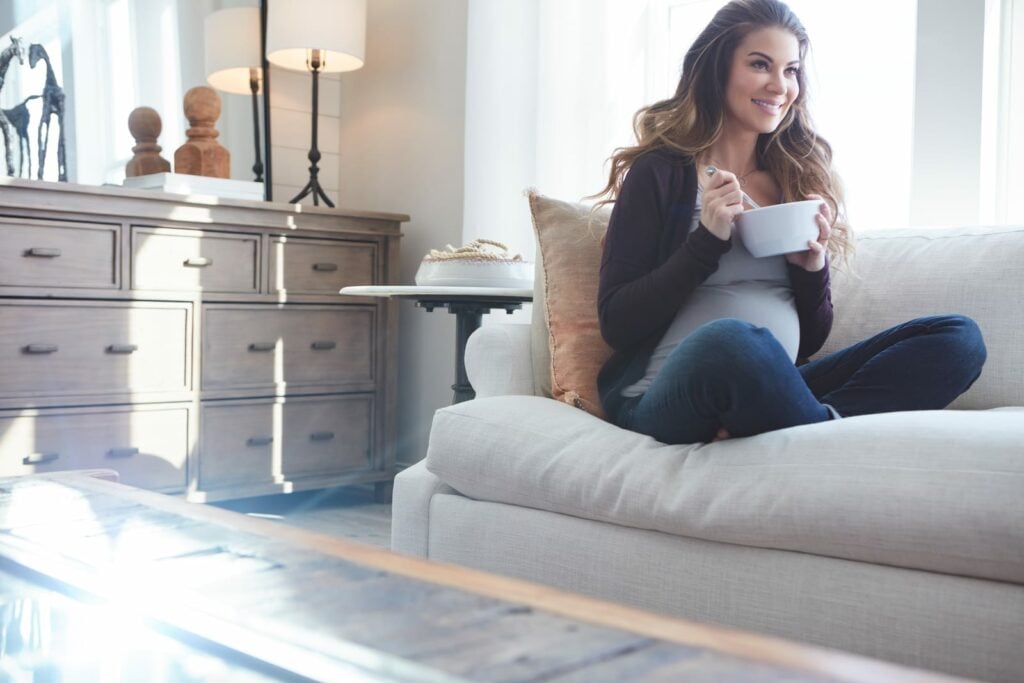 A woman sitting on a couch holding a bowl of breakfast while seeking ideas for how to get 30 grams of protein for breakfast.