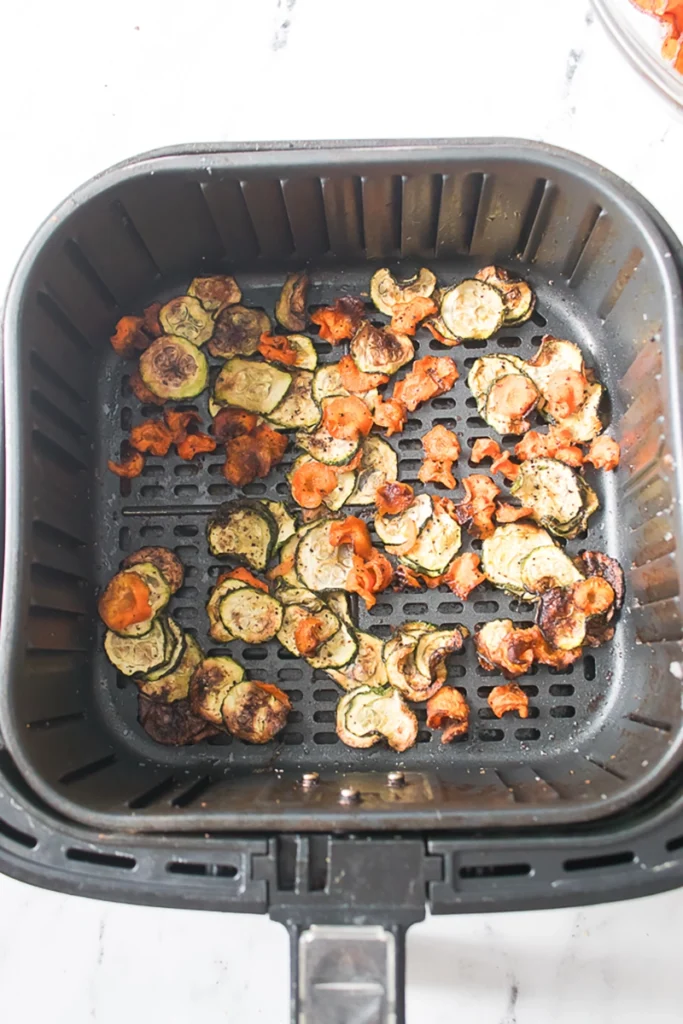 Slices of zucchini and carrots cooking in an air fryer basket, slightly browned and seasoned.