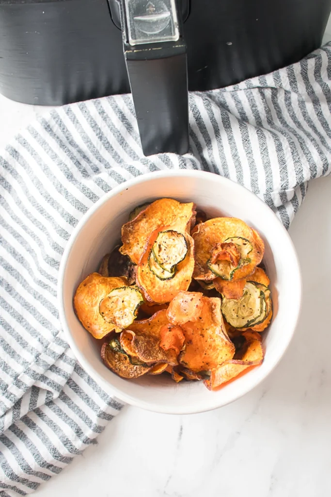 A white bowl filled with a mix of vegetable chips sits on a striped cloth next to an air fryer.