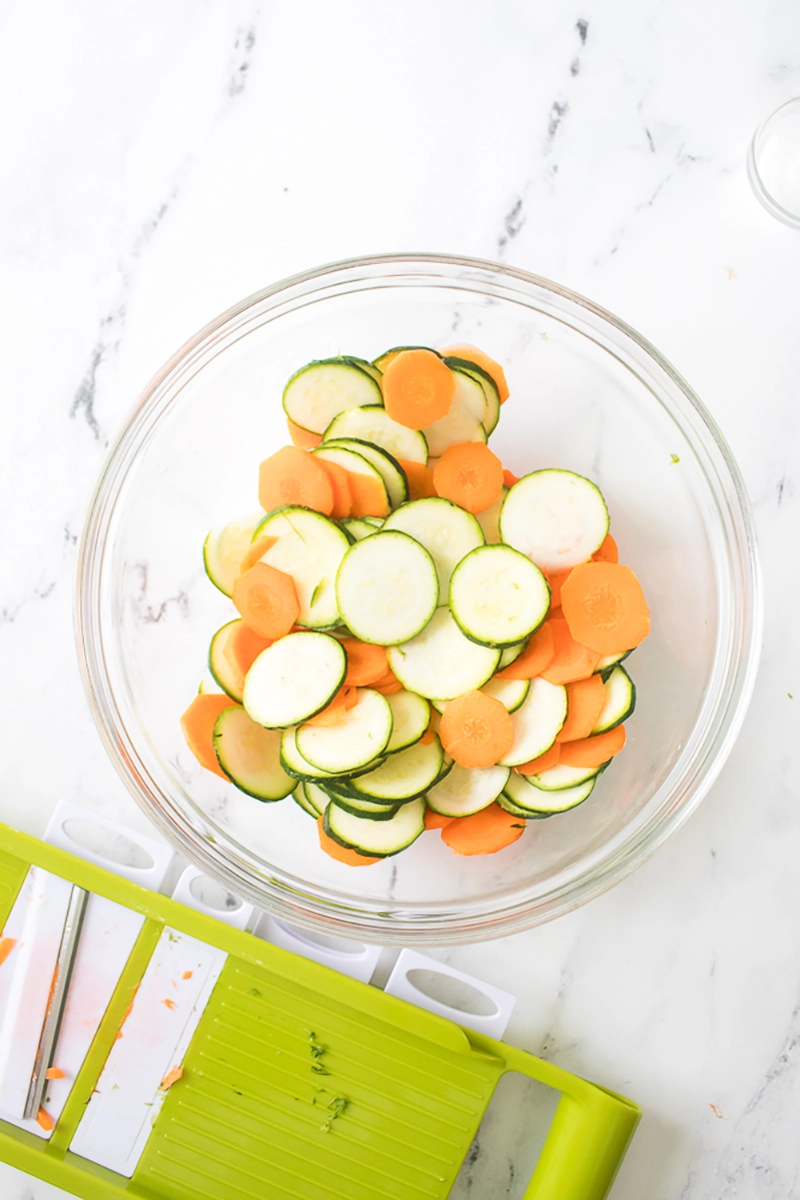 A glass bowl filled with sliced zucchini and carrots on a white marble surface, next to a green and white mandoline slicer.
