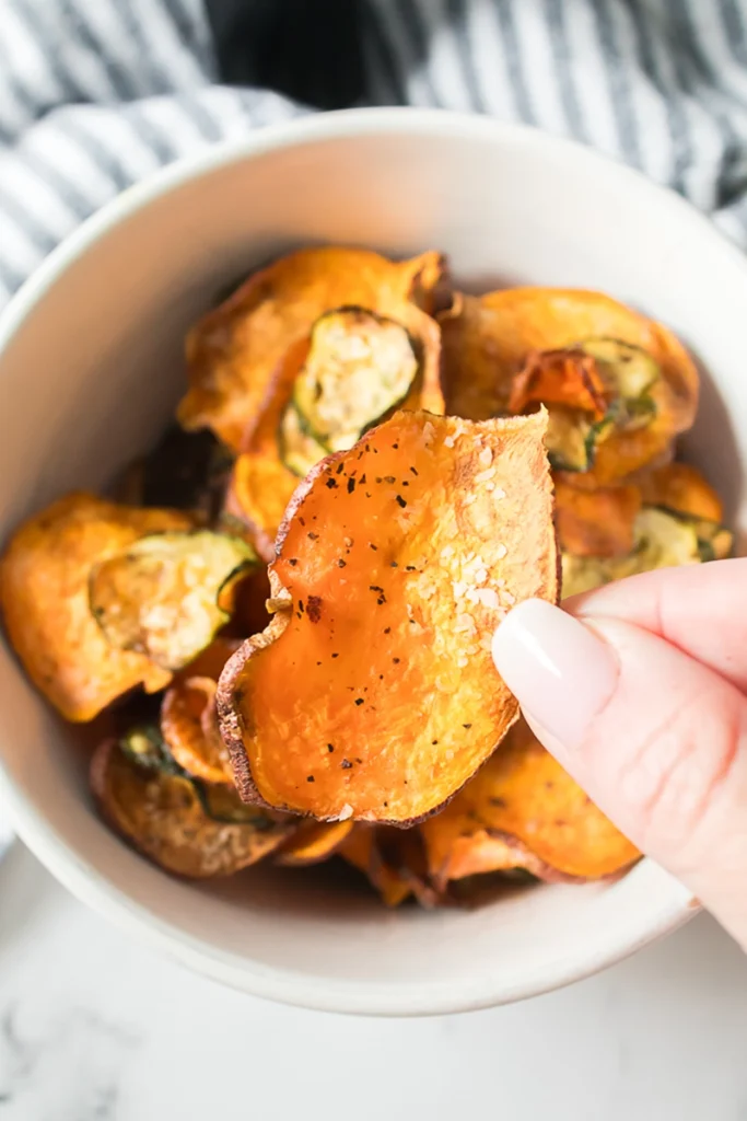 A hand holding a seasoned sweet potato chip over a bowl filled with similar chips.