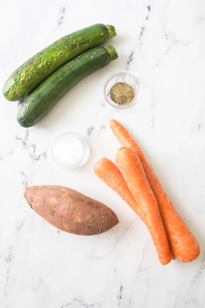 Two zucchinis, three carrots, one sweet potato, and small bowls of salt and pepper are arranged on a white marble surface.