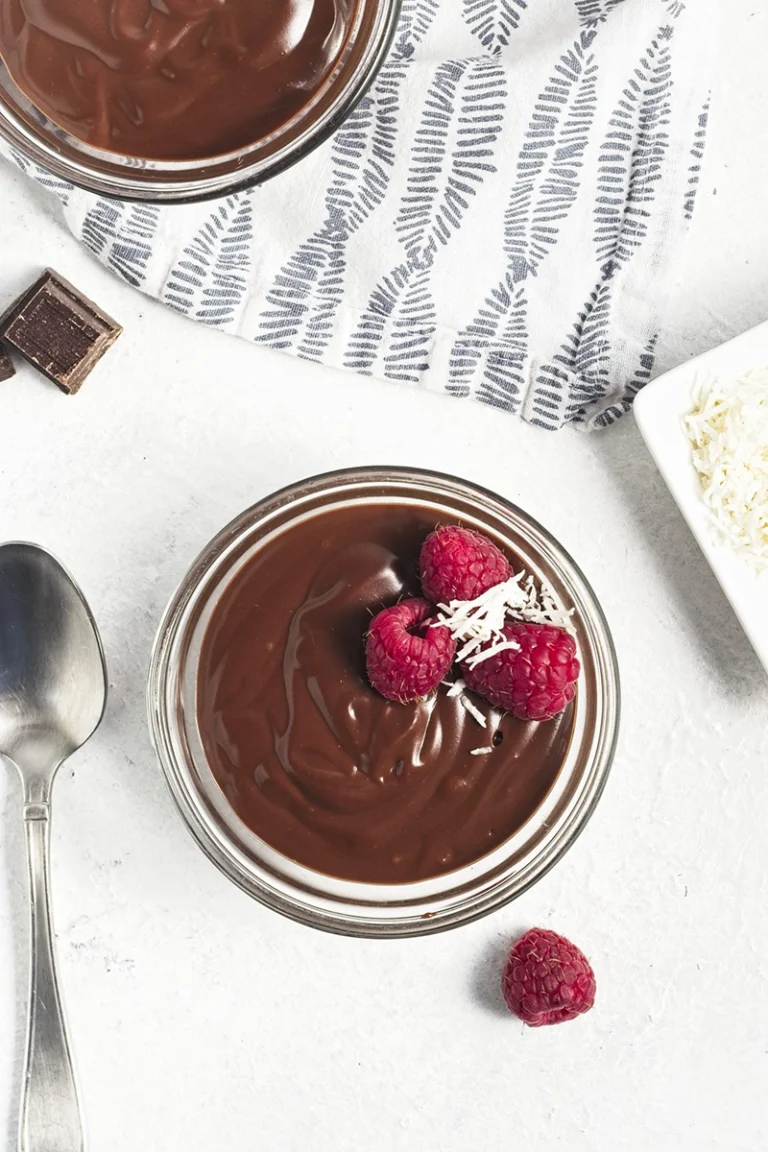 A glass bowl filled with chocolate pudding is topped with raspberries and coconut flakes, resting on a white surface next to a spoon, chocolate pieces, and a patterned cloth—an easy recipe perfect for two.