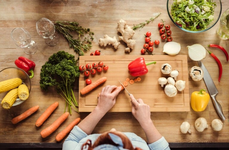 A person, perhaps seeking cooking for one advice, slices carrots on a wooden cutting board surrounded by various vegetables and herbs, including peppers, cherry tomatoes, garlic, onions, and corn, on a kitchen counter.