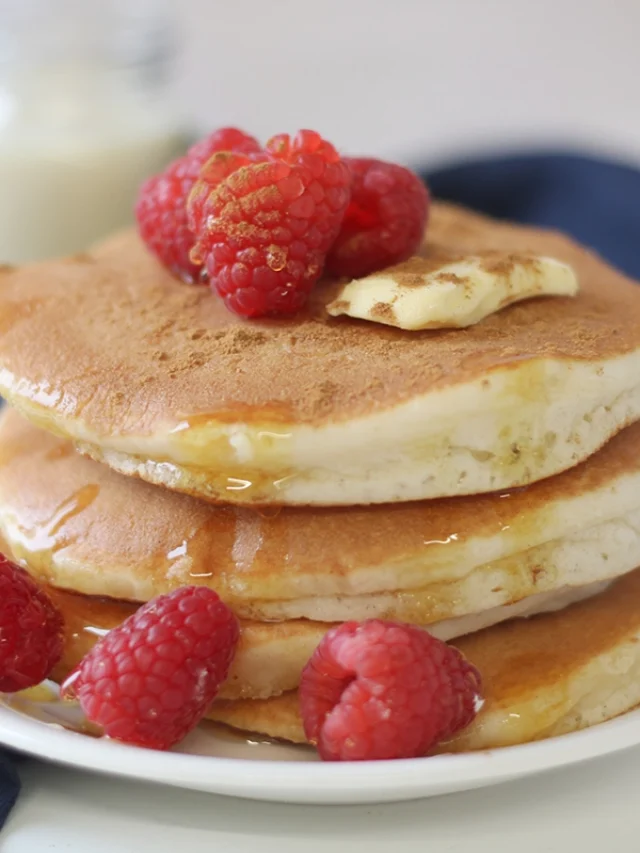 A single serving delight of gluten-free pancakes topped with fresh raspberries and a pat of butter, all drizzled with syrup. Additional raspberries adorn the plate.