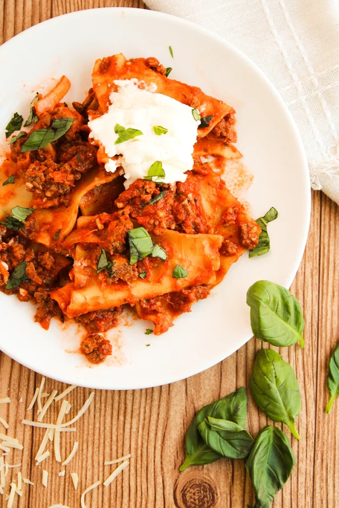 A skillet lasagna, rich with meat sauce and crowned with cheese and basil, rests invitingly on a wooden table, surrounded by scattered basil leaves and cheese shreds.