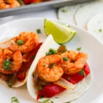 Two shrimp tacos with peppers and onions on a white plate, garnished with chopped herbs and lime wedges—these tasty 1 pan shrimp fajitas come together easily, with extra shrimp and vegetables in the background.