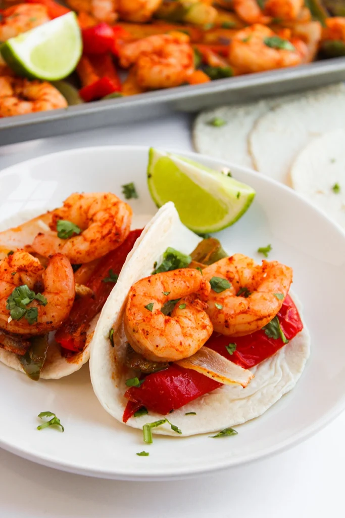 Two shrimp tacos with peppers and onions on a white plate, garnished with chopped herbs and lime wedges—these tasty 1 pan shrimp fajitas come together easily, with extra shrimp and vegetables in the background.