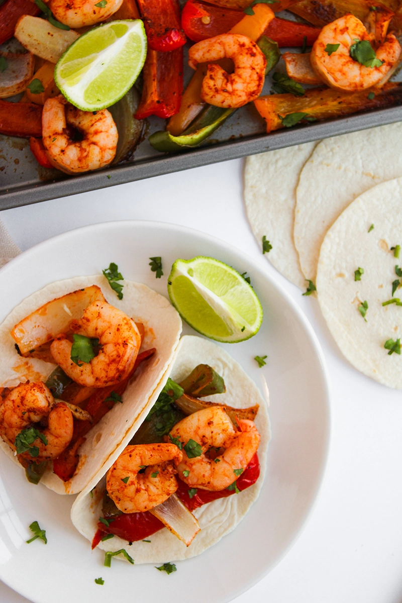Two sheetpan shrimp fajitas with vegetables on tortillas sit on a white plate, garnished with cilantro and lime, with extra tortillas and a tray of more 1 pan shrimp fajitas in the background.