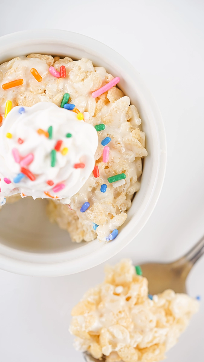 A single serving Rice Krispie treat sits in a bowl, topped with whipped cream and colorful sprinkles, with a bite taken out and a piece on a spoon beside this delightful individual dessert.