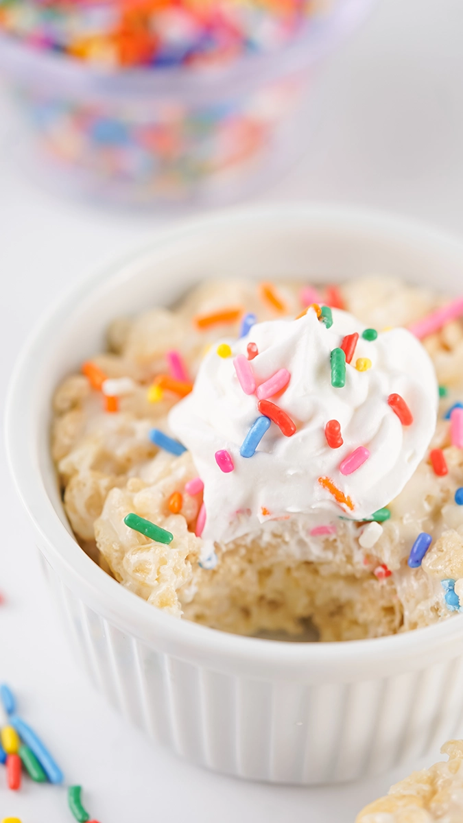 A small white ramekin filled with a single serving Rice Krispie Treat, topped with whipped cream and colorful sprinkles—an easy dessert that's as fun as it is delicious.