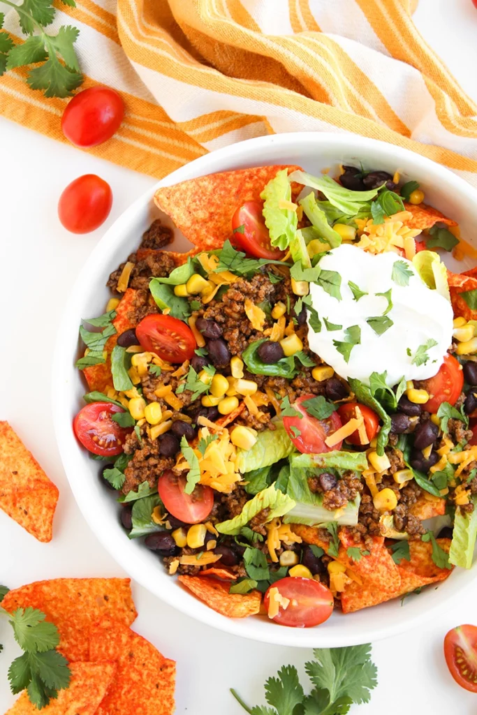 A bowl of doritos taco salad for one with ground beef, black beans, corn, lettuce, tomatoes, shredded cheese, tortilla chips, and a dollop of sour cream, garnished with cilantro.