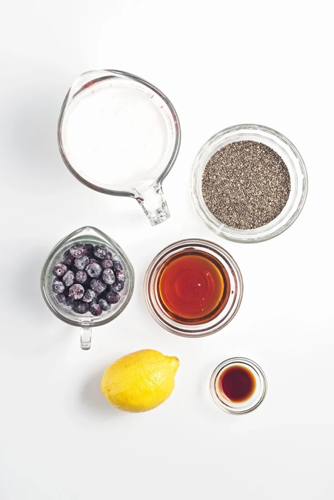 Six ingredients for blueberry lemon chia pudding on a white background: milk, chia seeds, blueberries, maple syrup, a lemon, and vanilla extract in clear containers.