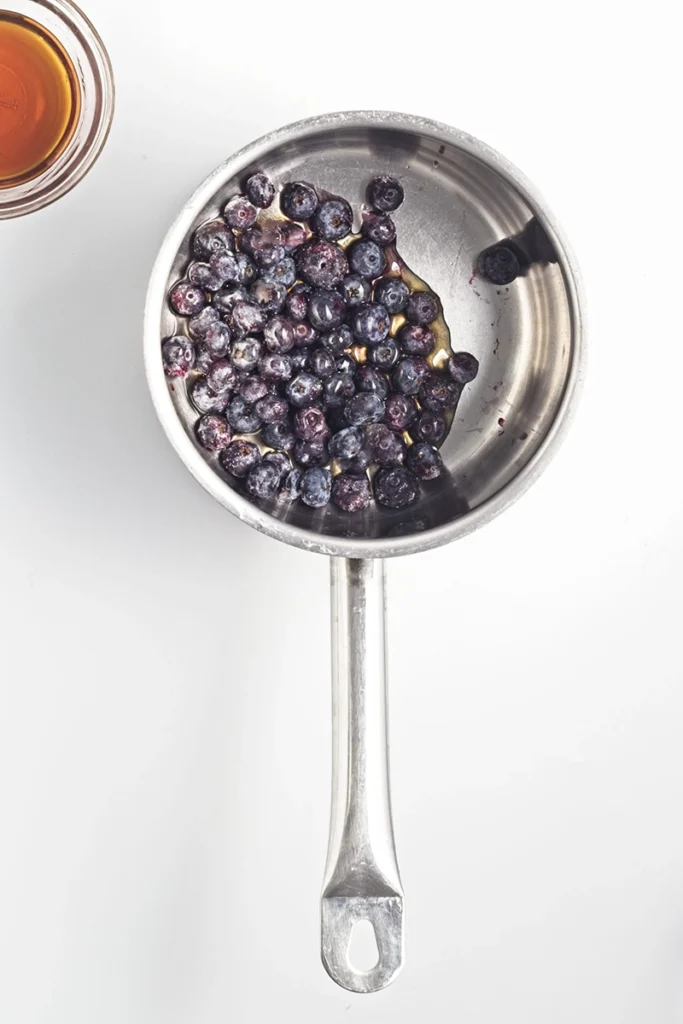 A stainless steel saucepan containing blueberries sits next to a glass of amber liquid, hinting at the start of a delicious blueberry chia seed pudding, all displayed on a white surface.