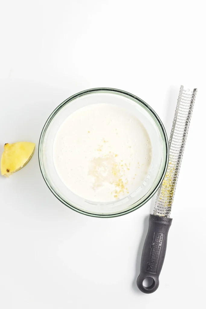 A glass bowl filled with creamy blueberry lemon chia pudding, sprinkled with lemon zest, sits beside a lemon wedge and a zester on a white surface.