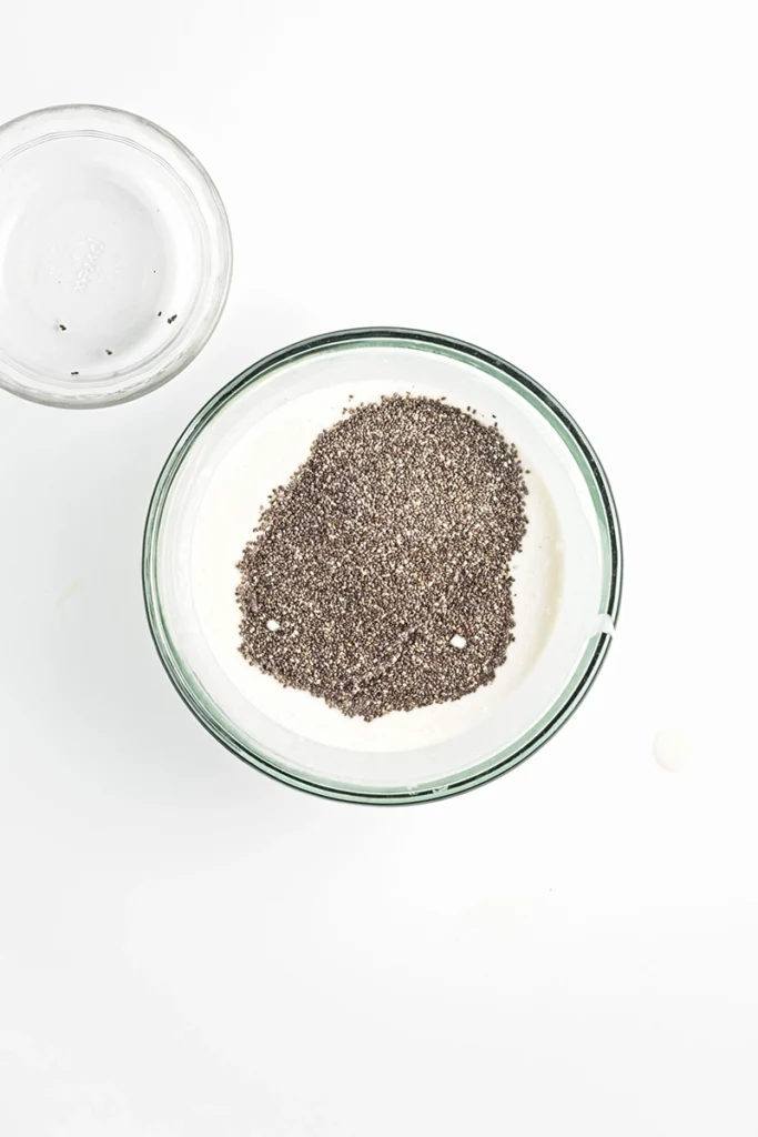 A glass bowl filled with creamy blueberry lemon chia pudding, topped with ground black pepper, sits beside a small empty glass bowl on a white surface.