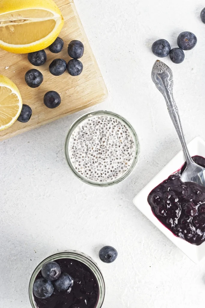 Overhead view of blueberry lemon chia pudding in a jar, accompanied by blueberry jam in a dish with a spoon, fresh blueberries, and lemon wedges on a cutting board.