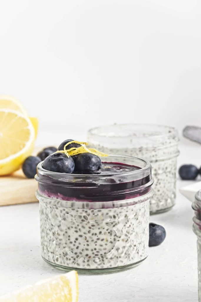 A glass jar of blueberry lemon chia pudding topped with blueberry sauce, fresh blueberries, and lemon zest, with lemons and more blueberries in the background.