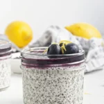 A glass jar filled with creamy chia seed pudding, topped with vibrant blueberry sauce, fresh blueberries, and lemon zest; lemons and a cloth are visible in the background.