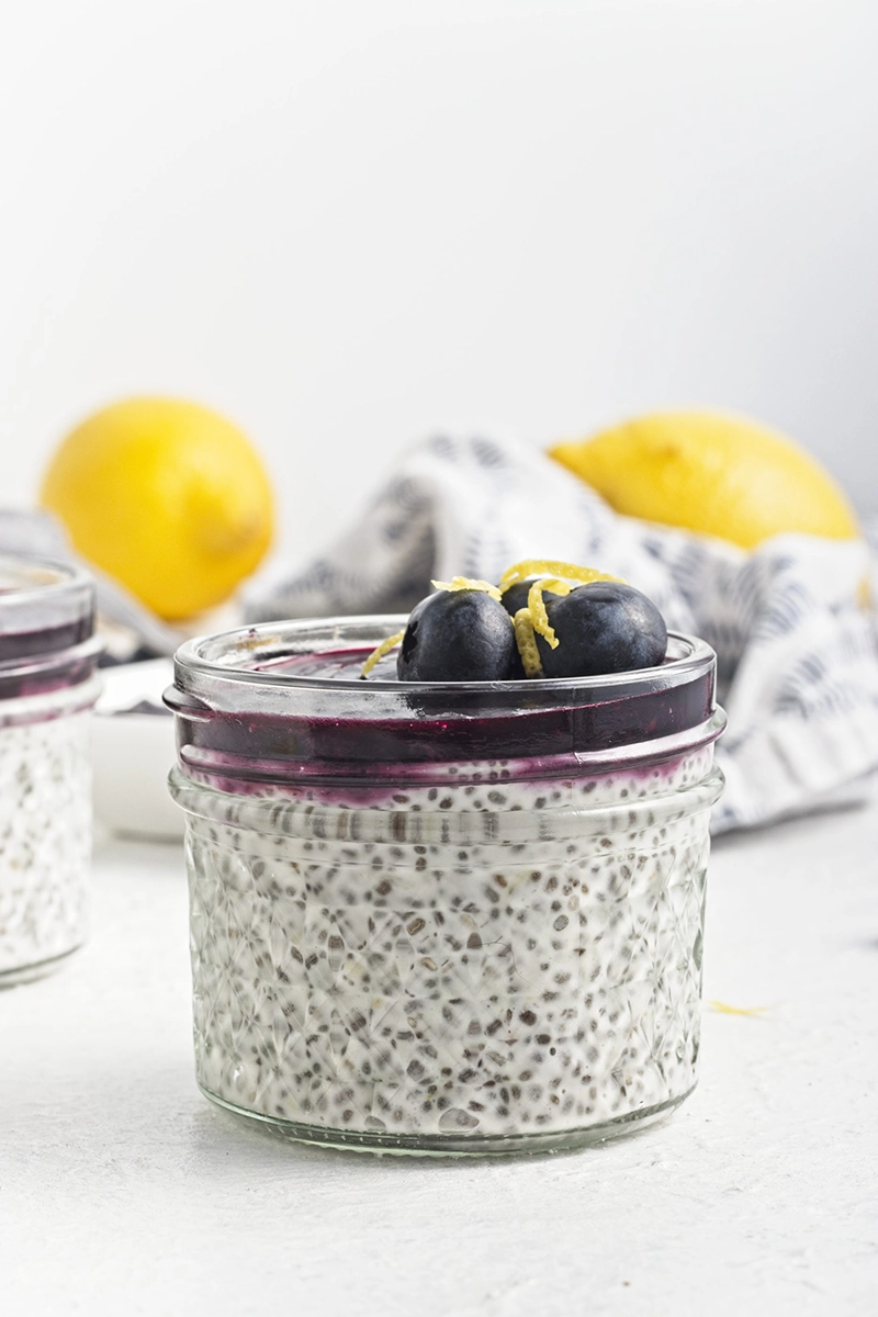 A glass jar filled with creamy chia seed pudding, topped with vibrant blueberry sauce, fresh blueberries, and lemon zest; lemons and a cloth are visible in the background.