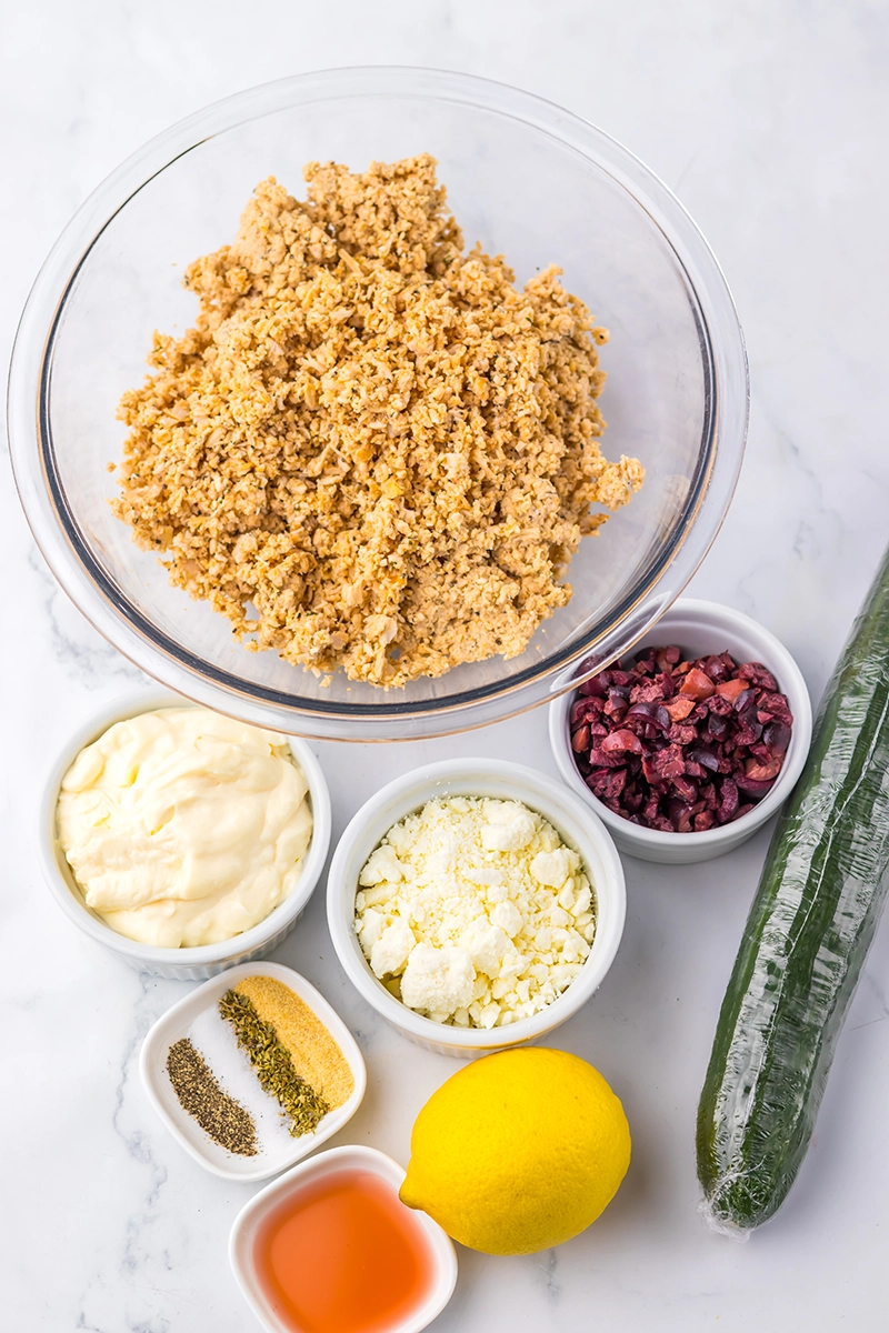 A glass bowl with crumbled chickpeas and small bowls containing mayo, dried cranberries, feta cheese, spices, vinegar, a lemon, and a cucumber on a marble surface—perfect for making a Greek goddess chicken salad.
