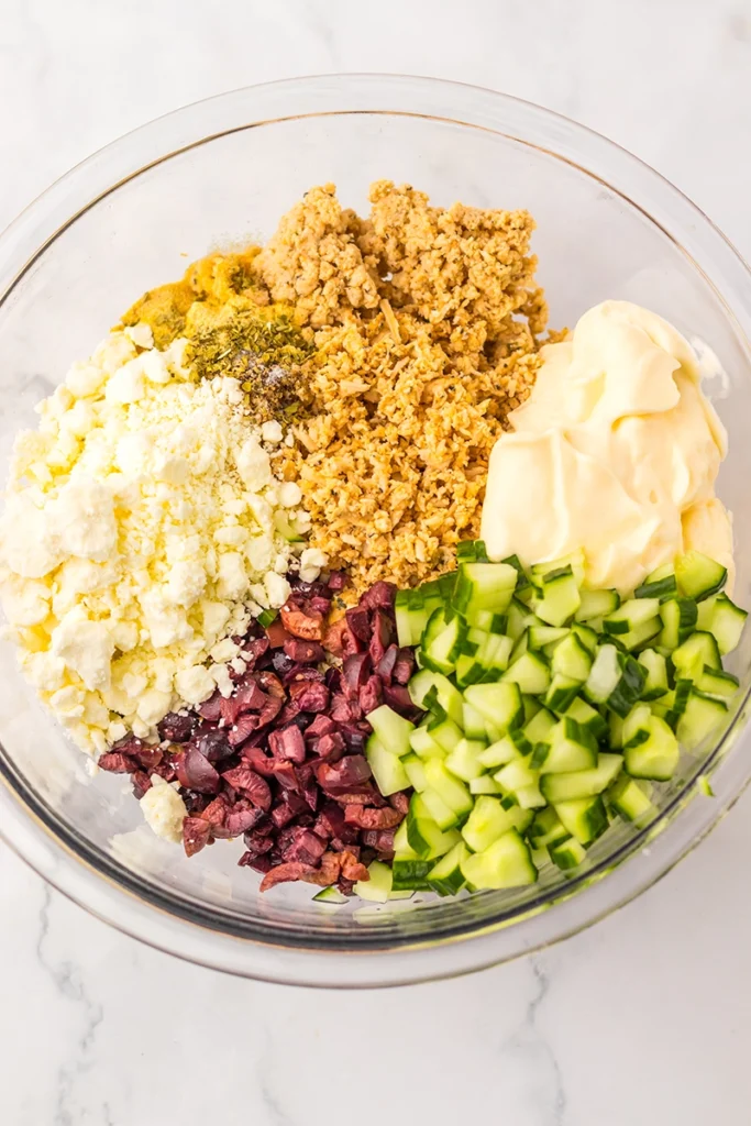A glass bowl containing feta cheese, cooked grains, mayonnaise, chopped cucumber, chopped olives, and seasonings—arranged in separate sections for a fresh greek goddess chicken salad twist.