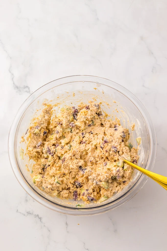 A glass bowl filled with a mixed batter reminiscent of a Greek goddess chicken salad, featuring visible pieces of vegetables and olives, with a yellow-handled spoon resting inside.