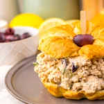 A croissant sandwich filled with Greek Goddess chicken salad and topped with a black olive, served on a gray plate beside a bowl of black olives.