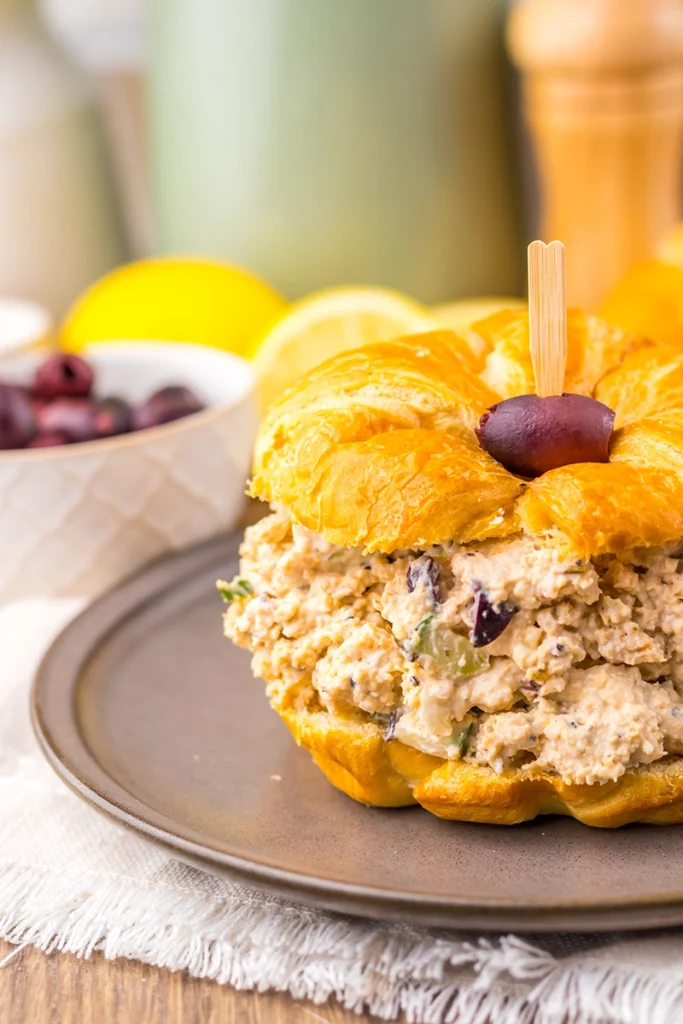 A croissant sandwich filled with Greek Goddess chicken salad and topped with a black olive, served on a gray plate beside a bowl of black olives.
