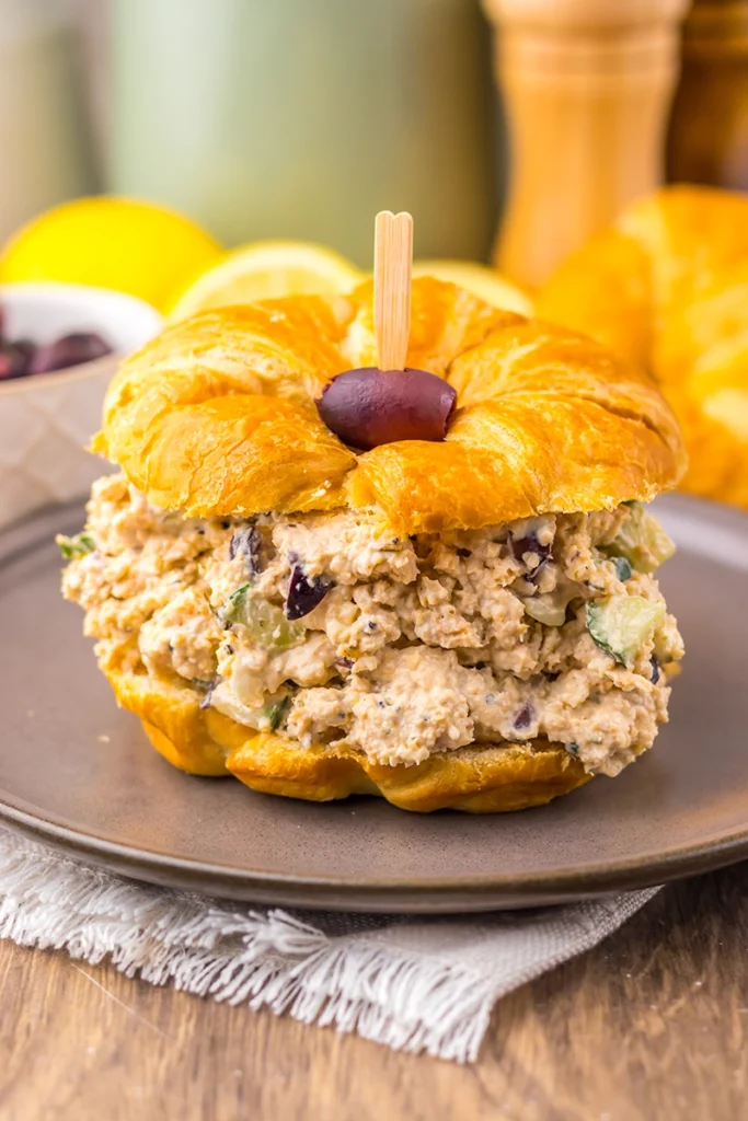 A croissant sandwich filled with Greek Goddess chicken salad and garnished with a grape half, served on a plate with a napkin underneath.