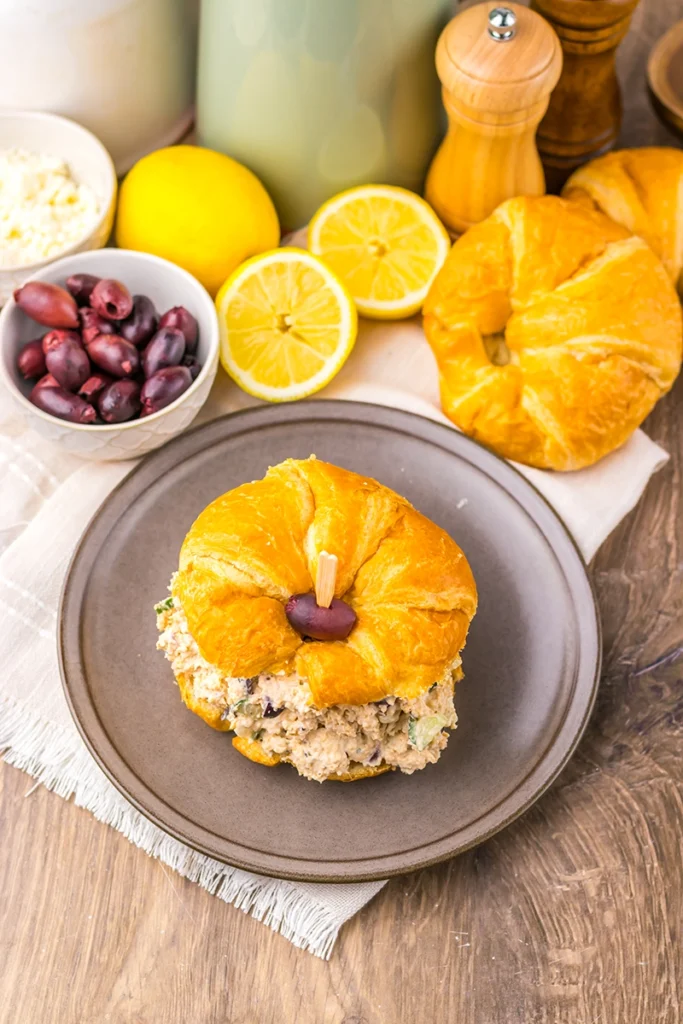 A croissant sandwich filled with a Greek goddess chicken salad and topped with a black olive sits on a plate. In the background are sliced lemons, a croissant, black olives, feta cheese, and seasoning shakers.