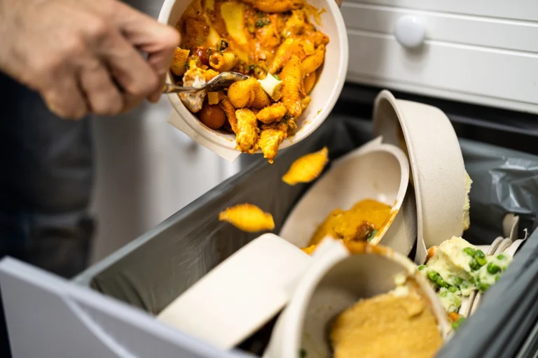 A hand dumps leftover food and disposable bowls into a kitchen trash bin filled with discarded meals—reminding us to use food waste tips and reduce food waste, especially when cooking for one.
