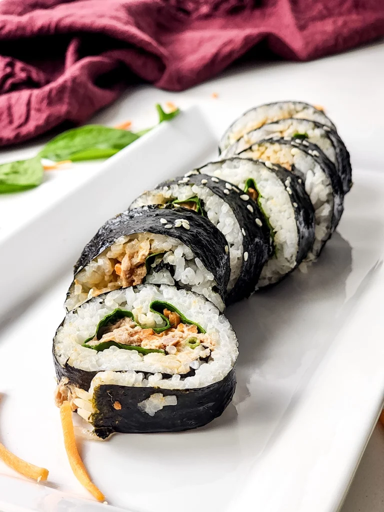 A row of sliced spicy tuna kimbap filled with rice, greens, and a brown filling is arranged on a white rectangular plate—perfect for trying your favorite spicy tuna kimbap recipe.