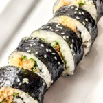 A close-up of sliced spicy tuna kimbap with seaweed, rice, flavorful filling, and sprinkled sesame seeds arranged on a white plate.