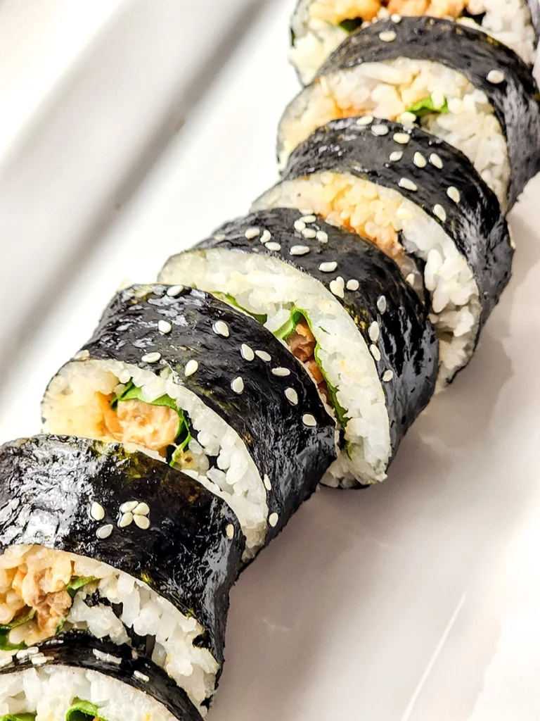 A close-up of sliced spicy tuna kimbap with seaweed, rice, flavorful filling, and sprinkled sesame seeds arranged on a white plate.