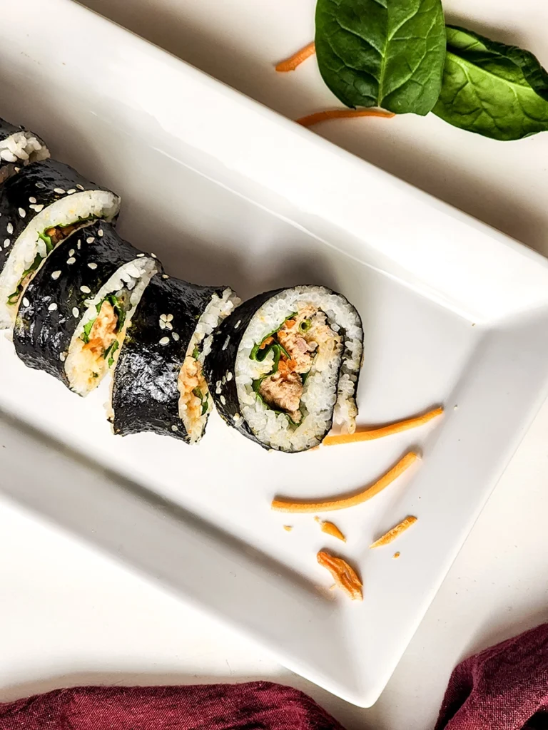 Sliced sushi rolls filled with rice, greens, and a creamy filling are arranged on a rectangular white plate, perfect for a spicy tuna kimbap recipe, with spinach leaves and shredded cheese nearby.