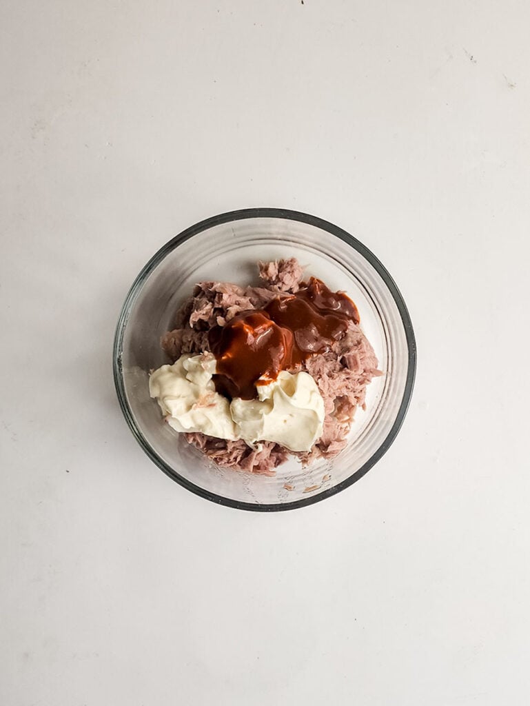 A clear glass bowl containing shredded meat topped with mayonnaise and barbecue sauce, inspired by spicy tuna kimbap, placed on a white surface.