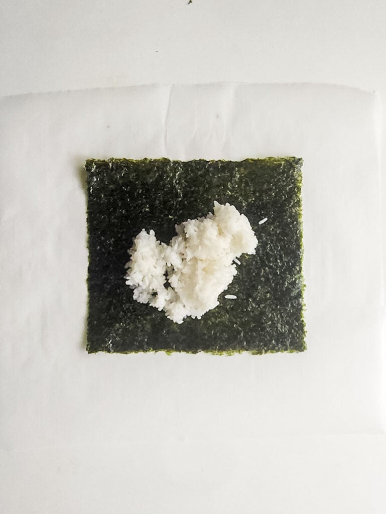 A sheet of nori seaweed on white parchment paper with a small pile of white rice in the center, ready to be filled as part of a spicy tuna kimbap recipe.