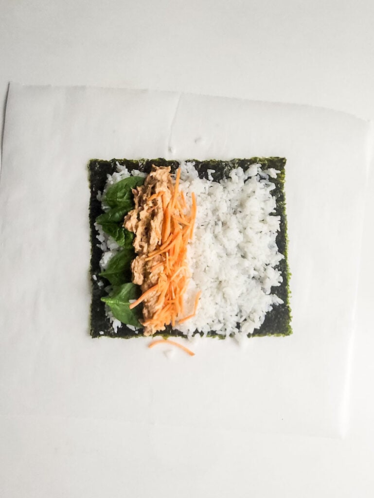 A sheet of seaweed topped with white rice, spinach leaves, shredded carrot, and a zesty tuna mixture—perfect for rolling your own spicy tuna kimbap at home.