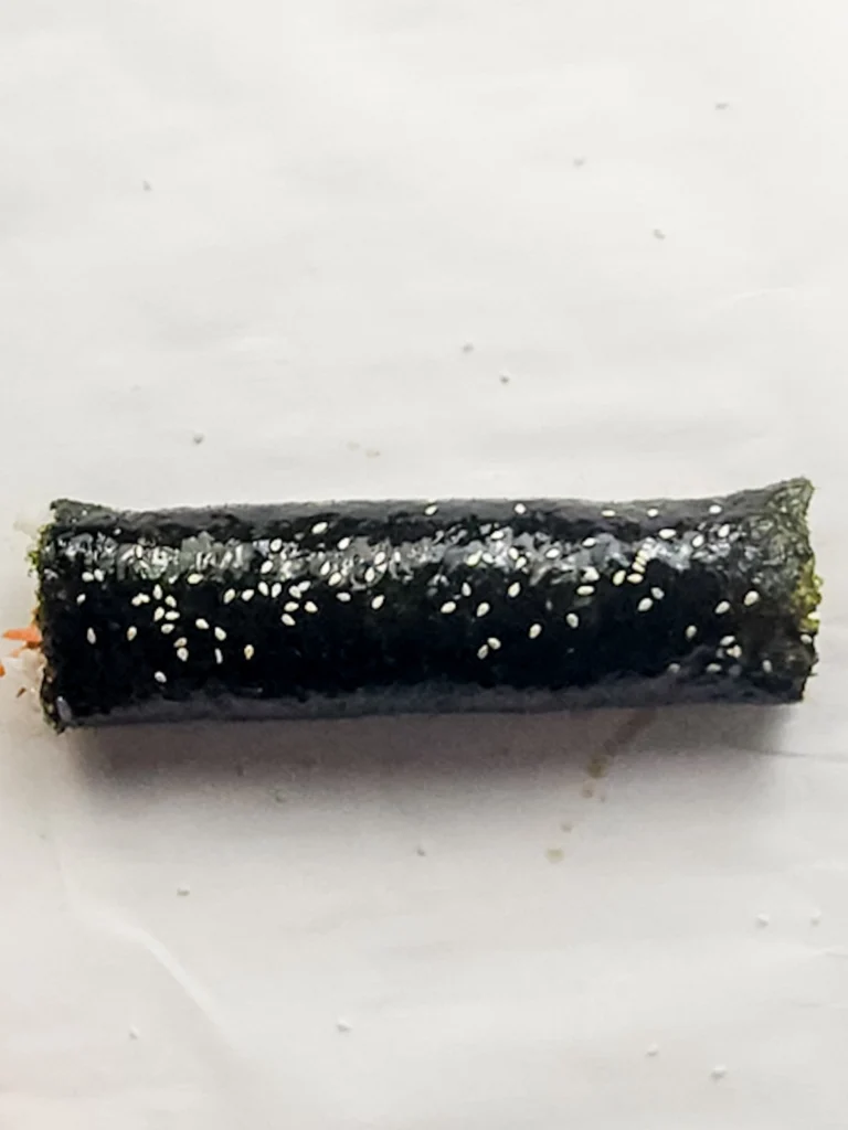 A sushi roll wrapped in seaweed and sprinkled with white sesame seeds on a white surface, perfect for trying your own spicy tuna kimbap recipe.