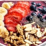 Enjoy a delicious single serving acai bowl topped with banana slices, strawberries, blueberries, chia seeds, granola, and sliced almonds, beautifully presented in a white bowl.