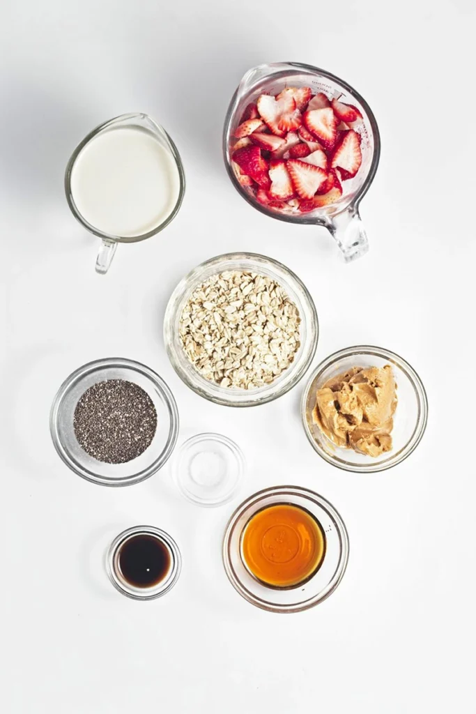 Seven clear bowls and measuring cups containing all the ingredients for peanut butter and jam overnight oats—milk, sliced strawberries, oats, chia seeds, peanut butter, vanilla extract, maple syrup, and salt—arranged on a white background.