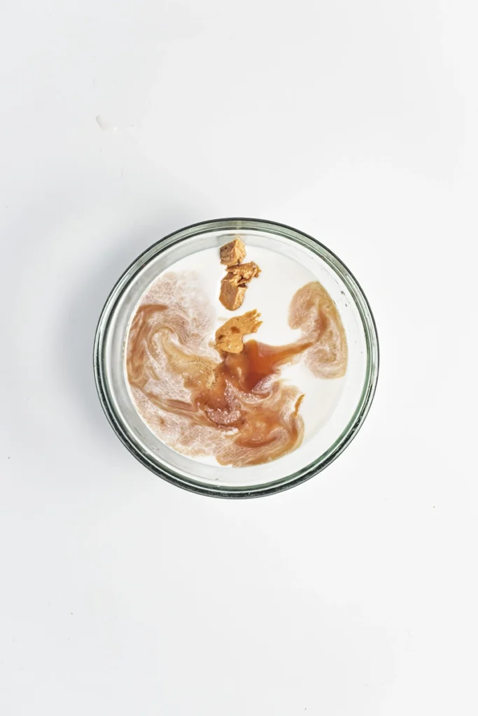 A glass bowl containing milk with a swirl of brown liquid and a few chunks floating on the surface, resembling peanut butter and jelly overnight oats, viewed from above on a white background.
