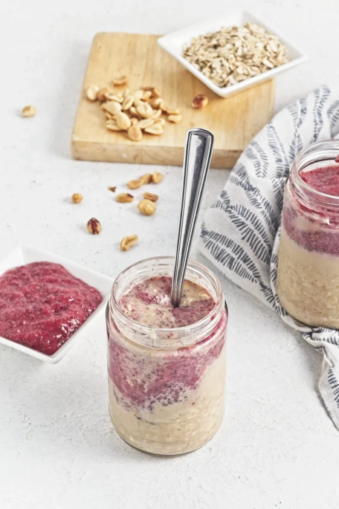 A jar of peanut butter and jelly overnight oats topped with a layer of red fruit puree, with a spoon inside. Peanuts and oats are on a wooden board in the background.