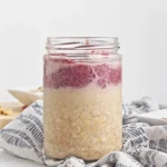 A glass jar filled with peanut butter and jelly overnight oats, featuring oatmeal on the bottom and a layer of red fruit puree on top, placed on a patterned cloth.