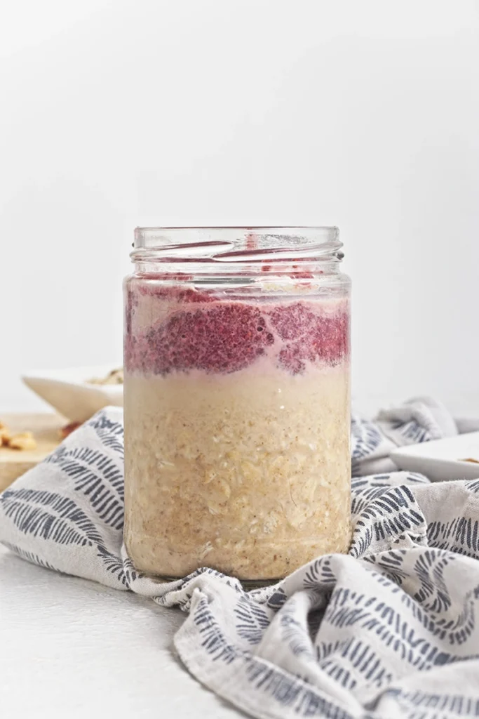 A glass jar filled with peanut butter and jelly overnight oats, featuring oatmeal on the bottom and a layer of red fruit puree on top, placed on a patterned cloth.