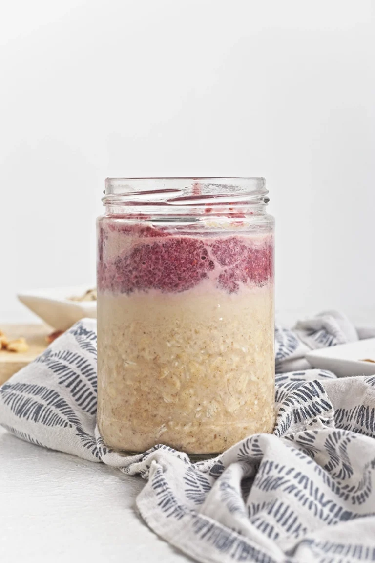 A glass jar filled with peanut butter and jelly overnight oats, featuring oatmeal on the bottom and a layer of red fruit puree on top, placed on a patterned cloth.